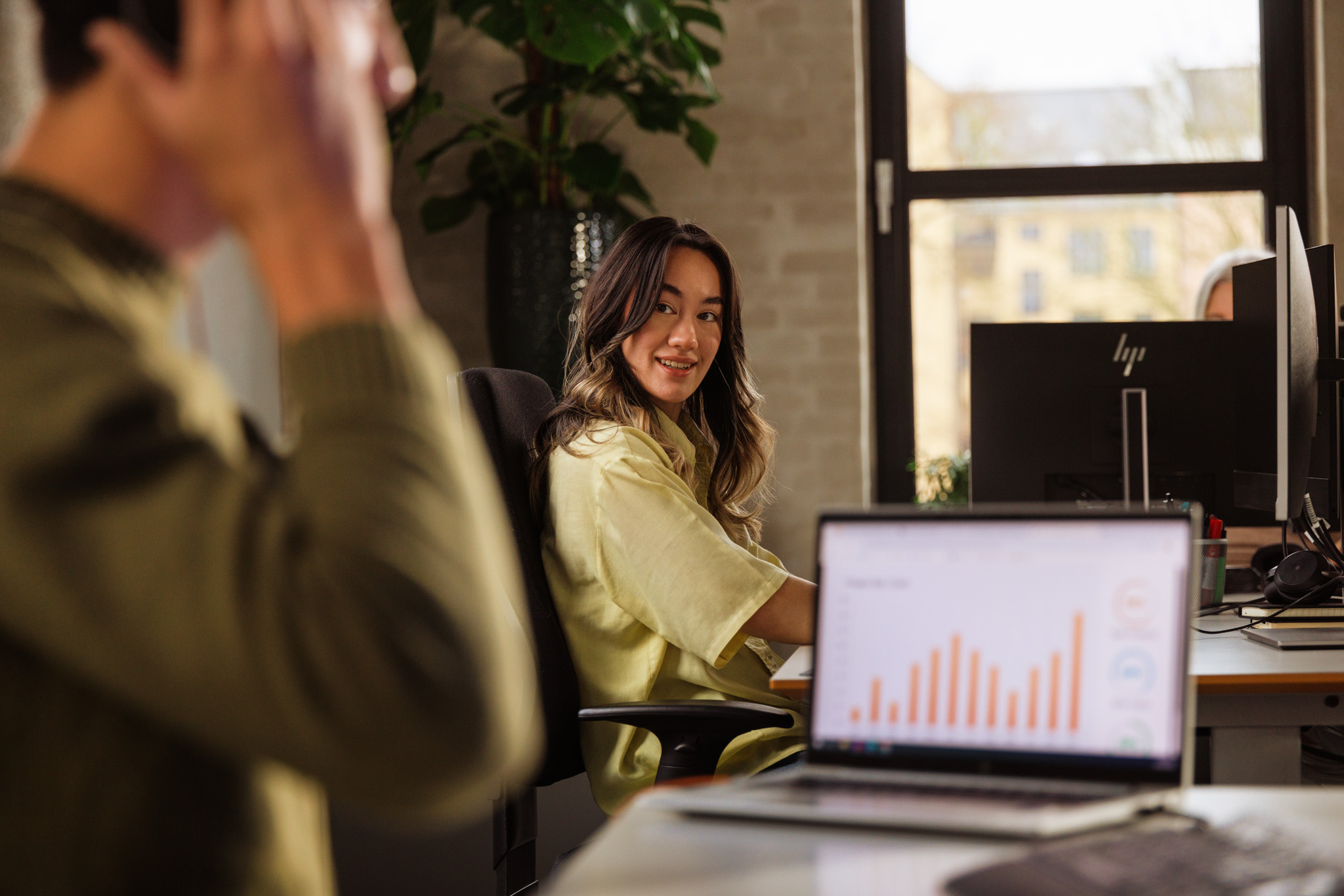 Woman sitting and talking at desk looking at her male coworker who is at his desk joining a virtual meeting