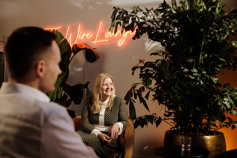 A woman sitting in an office surrounded by house plants looking out the window with a neon sign behind her