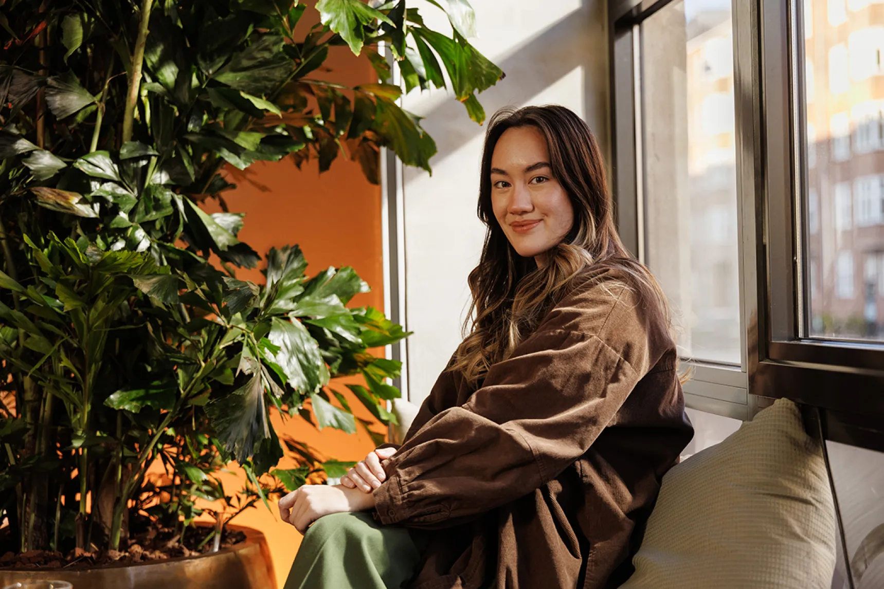 Woman sitting and smiling at the camera, surrounded by a large office plant and warm orange lighting.