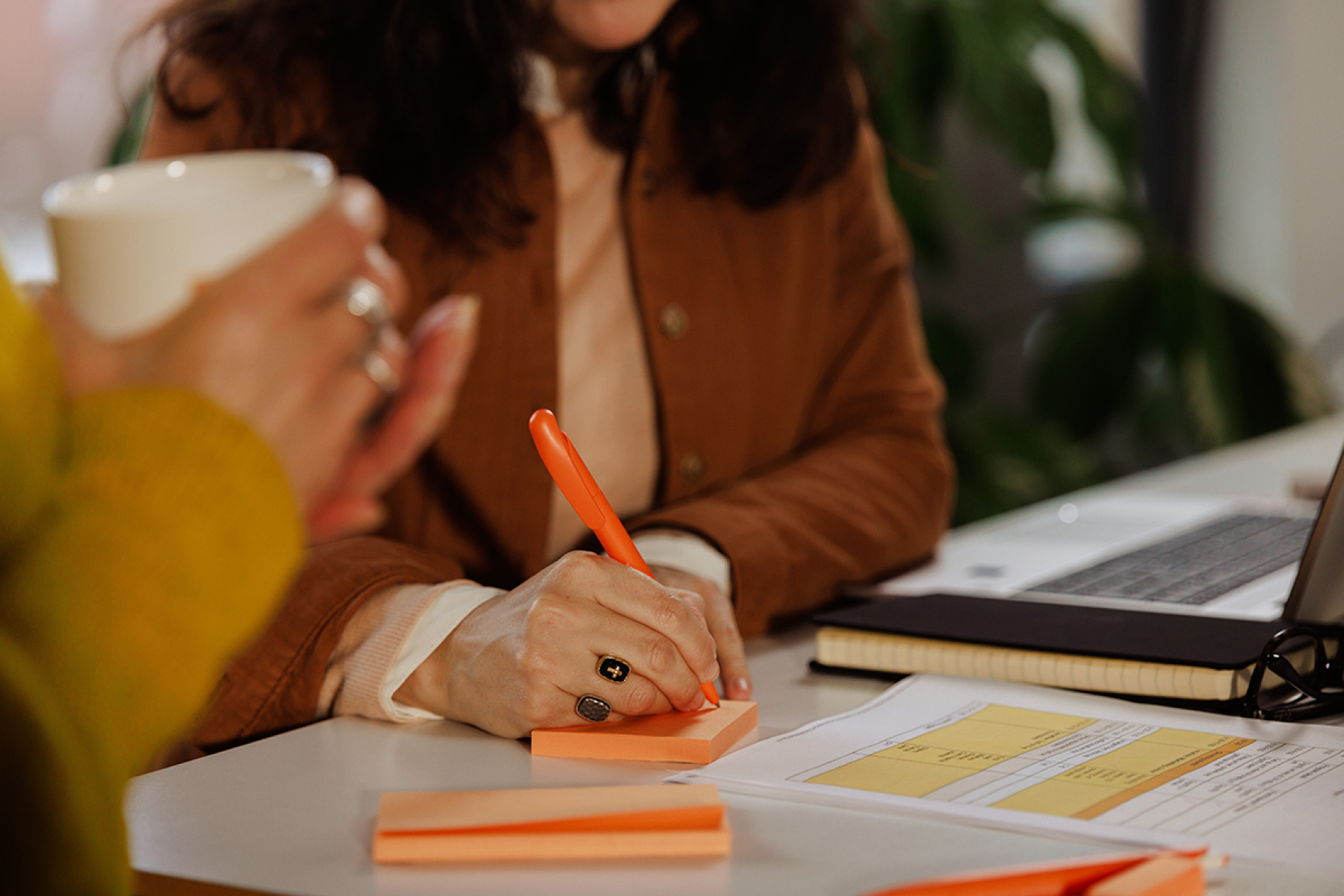 Woman writing with an orange pen on an orange sticky note next to her colleague holding a white coffee cup
