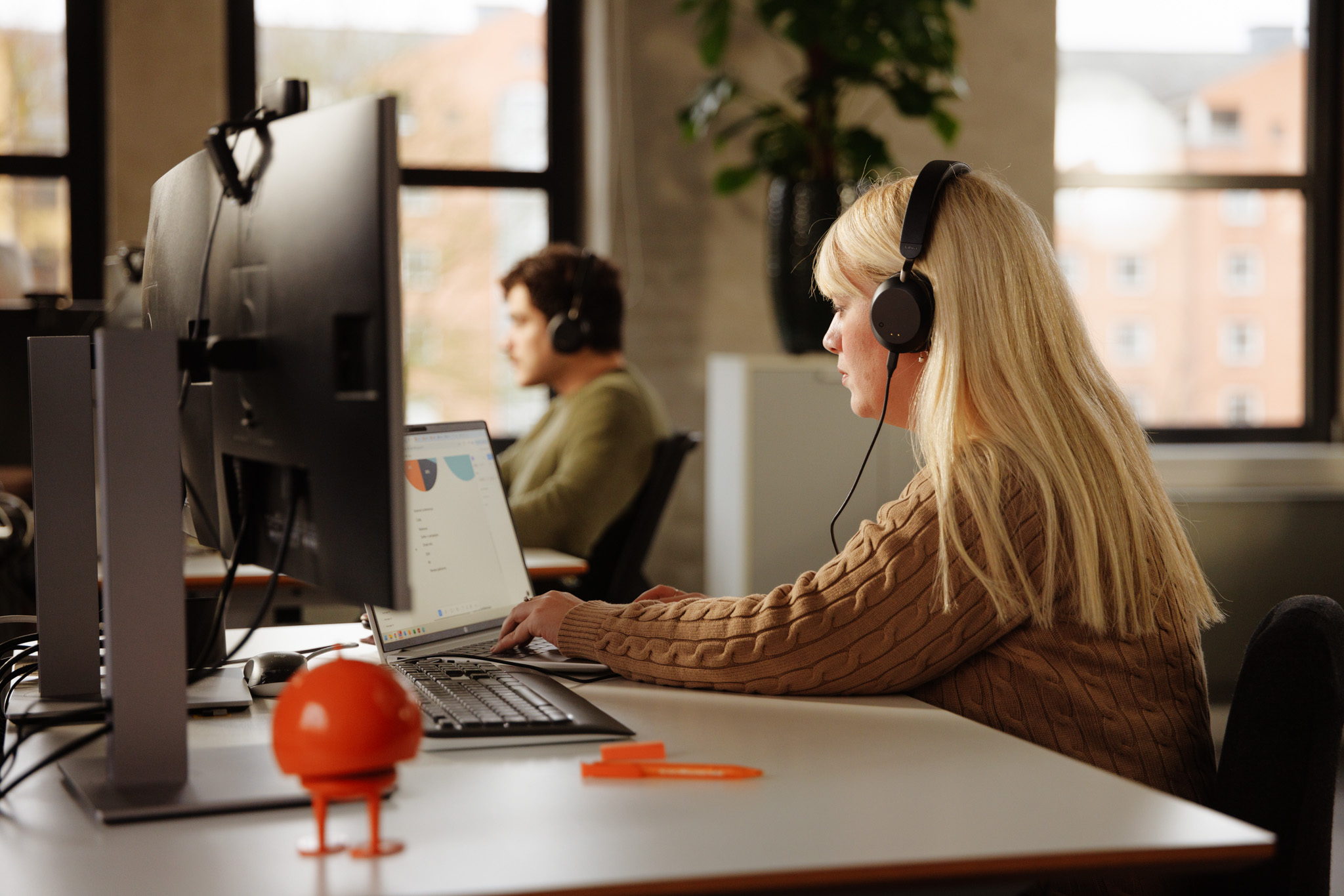 Woman sitting at desk looking at computer with data on screen while typing and wearing headphones 